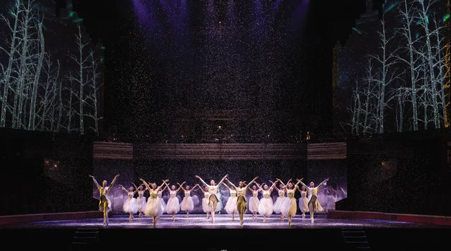 A group of ballet dancers in white snowflake costumes stand on a snow covered stage. 