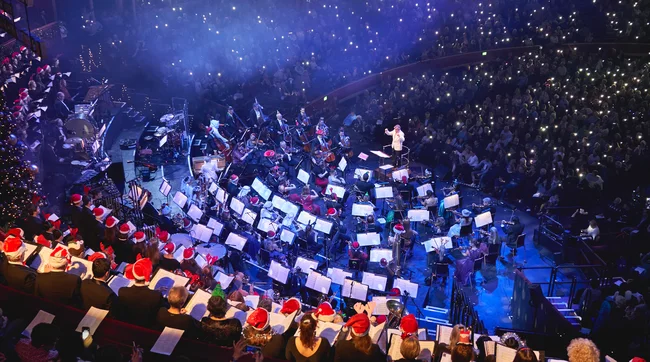 A large choir and orchestra sit on a stage with a conductor in front of them, wearing santa hats. 