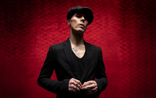 VV dressed in all black including a black hat, holding his hands together in front of him standing in front of a red background