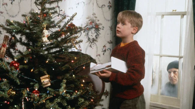 Macaulay Culkin as Kevin McCallister in Home Alone. He stands in a red jumper putting decorations on a Christmas tree.