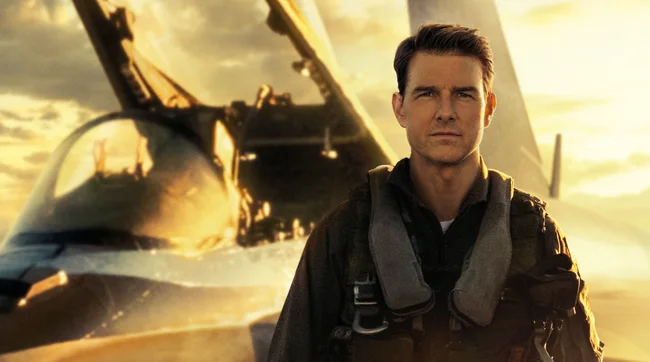 Tom Cruise in Top Gun face on to the camera in Air Force uniform with plane / clouds behind him. 