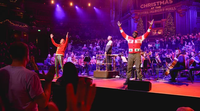 My Great Orchestral Adventure 2023, two presents with hands in the air in red Christmas jumpers in front of an orchestra