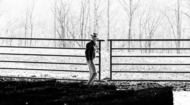 A man in a cowboy hat and denim jacket stands next to a large metal farm gate. 